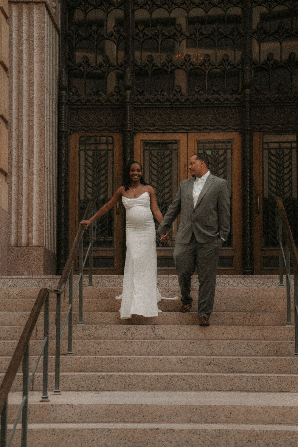 A bride and groom hold hands walking down stone steps before ornate bronze doors in a wedding portrait.