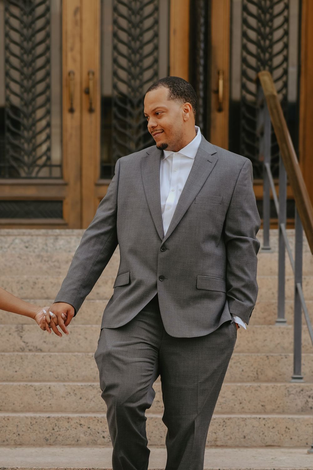 A well-dressed man in a gray suit holds hands while descending stone steps outside an elegant building.