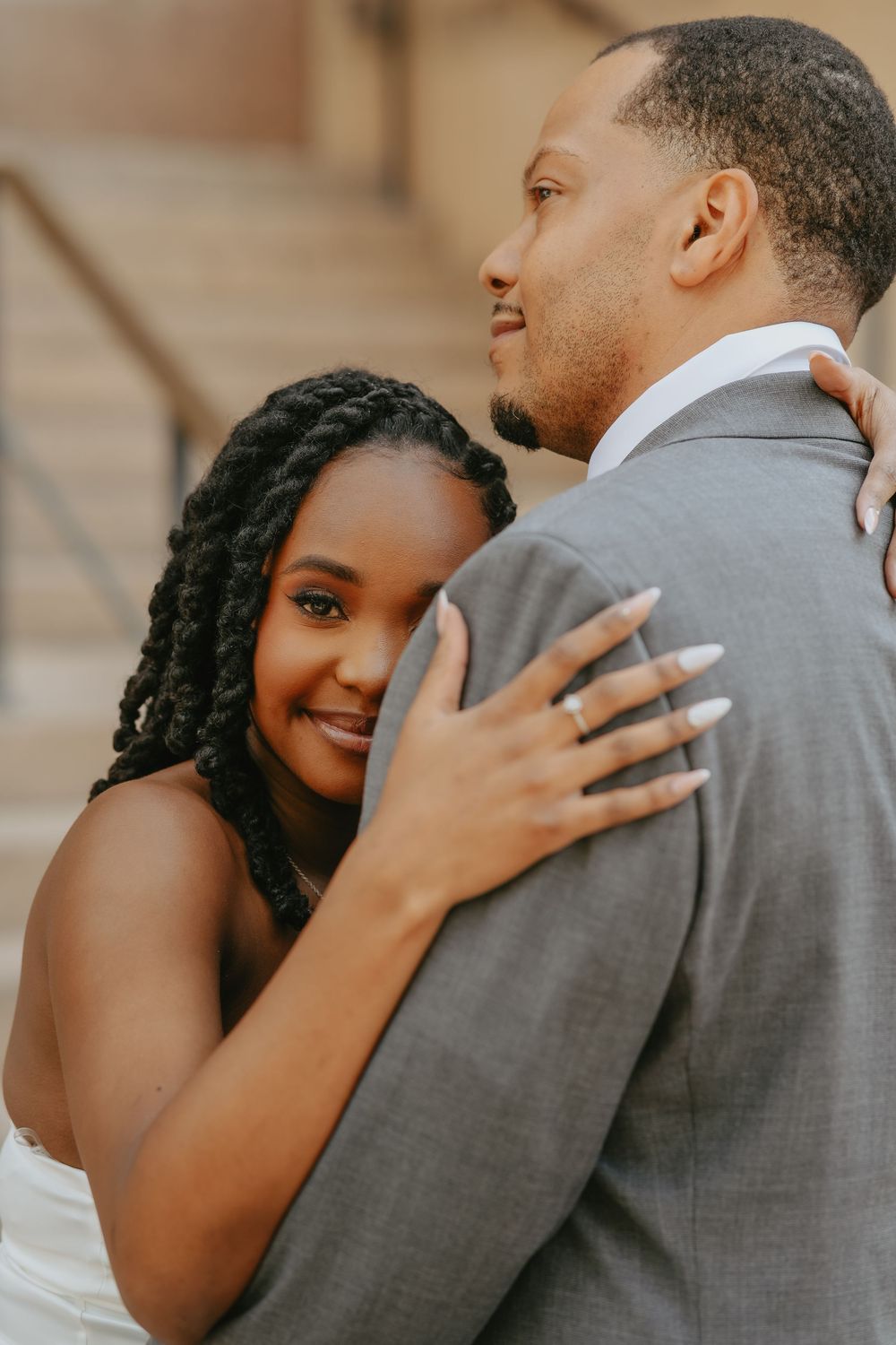 A bride with braided hair embraces a groom in a gray suit on stone steps in a romantic wedding portrait.