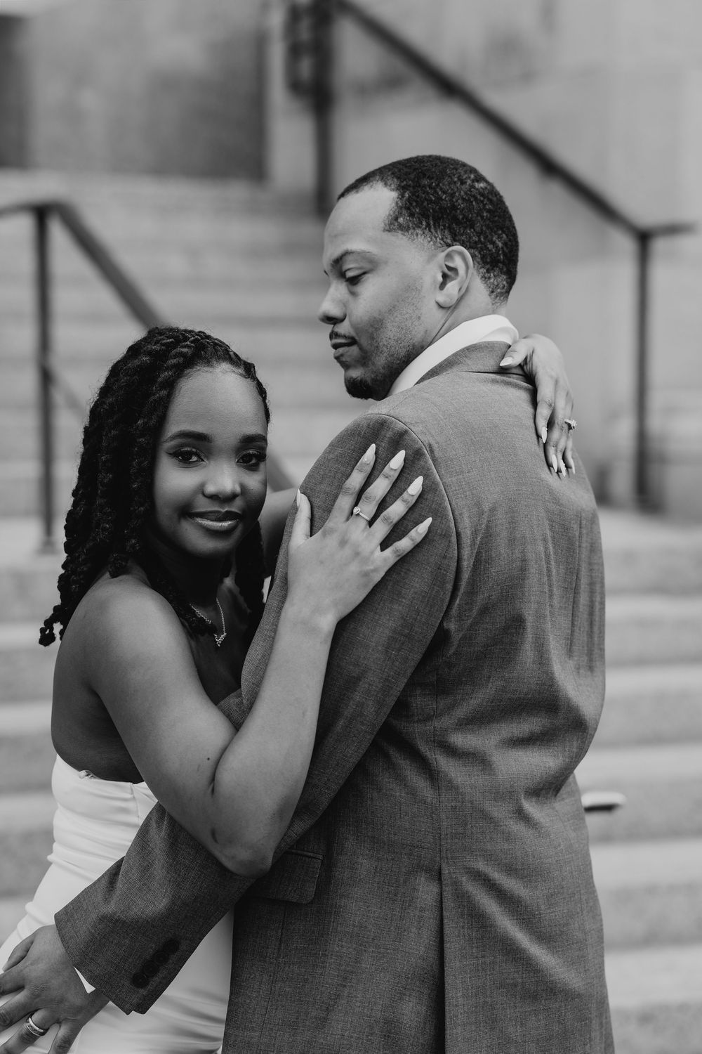 Black and white photo of a couple embracing on steps, woman smiling at camera while man looks down tenderly.
