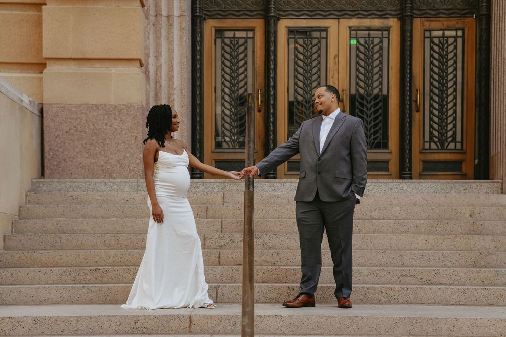 Bride in white gown and groom in gray suit hold hands on stone steps before ornate Art Deco building doors.