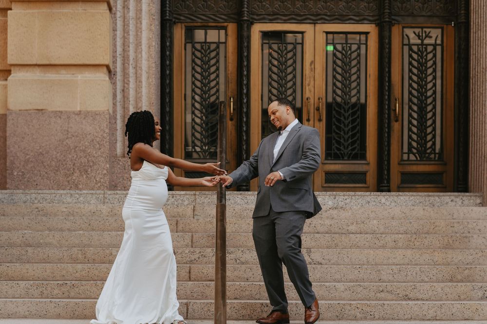 Bride in white gown and groom in gray suit hold hands on stone steps outside elegant building with ornate doors.