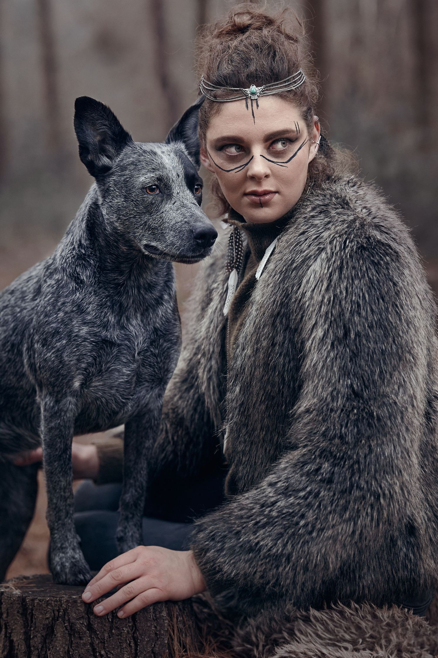 Woman in fur coat with tribal makeup and headdress poses with a gray dog in a misty forest setting.