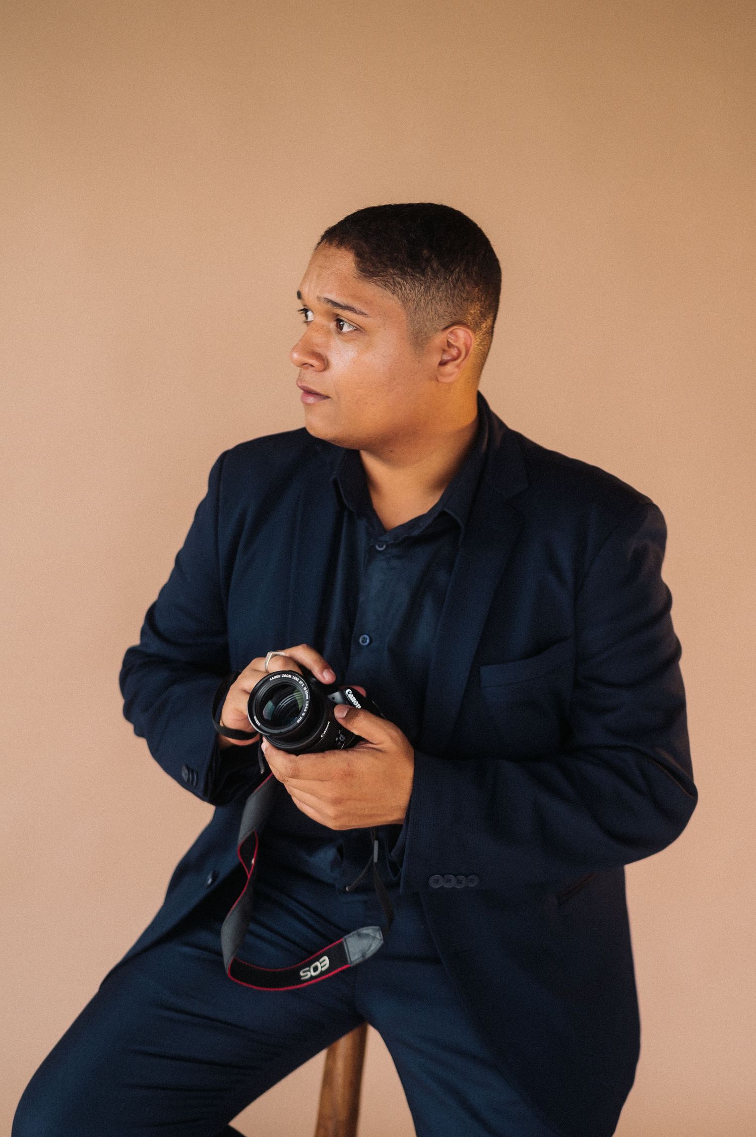 Young man in navy suit holding a DSLR camera while seated against a beige studio background.