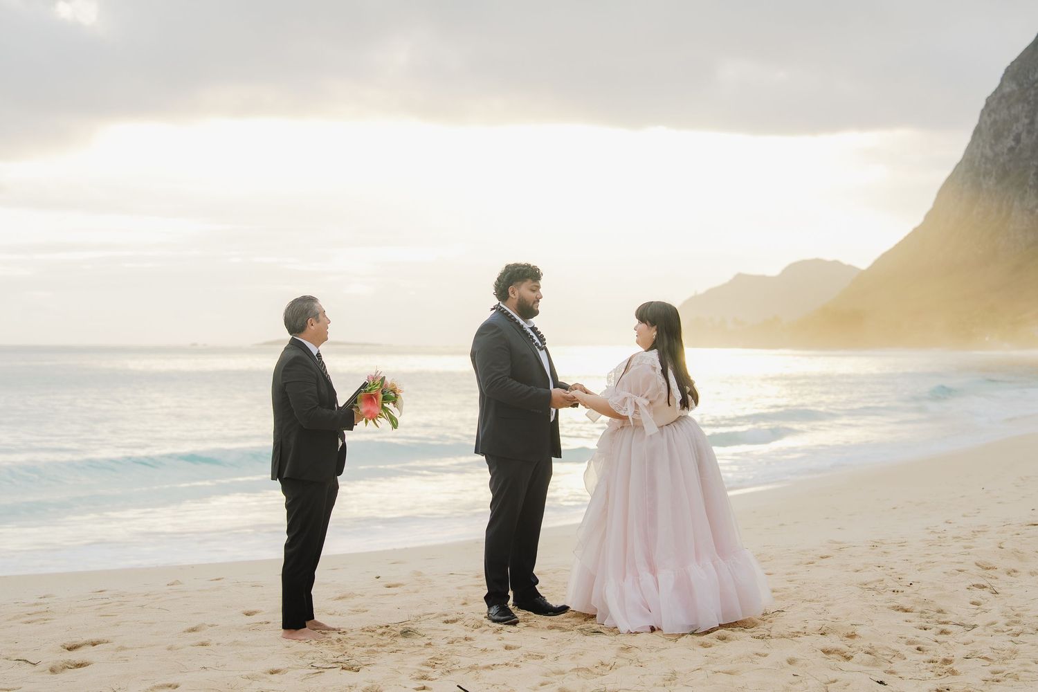 Couple exchanging vows on a sandy beach at sunset with an officiant, ocean waves and mountains in background.