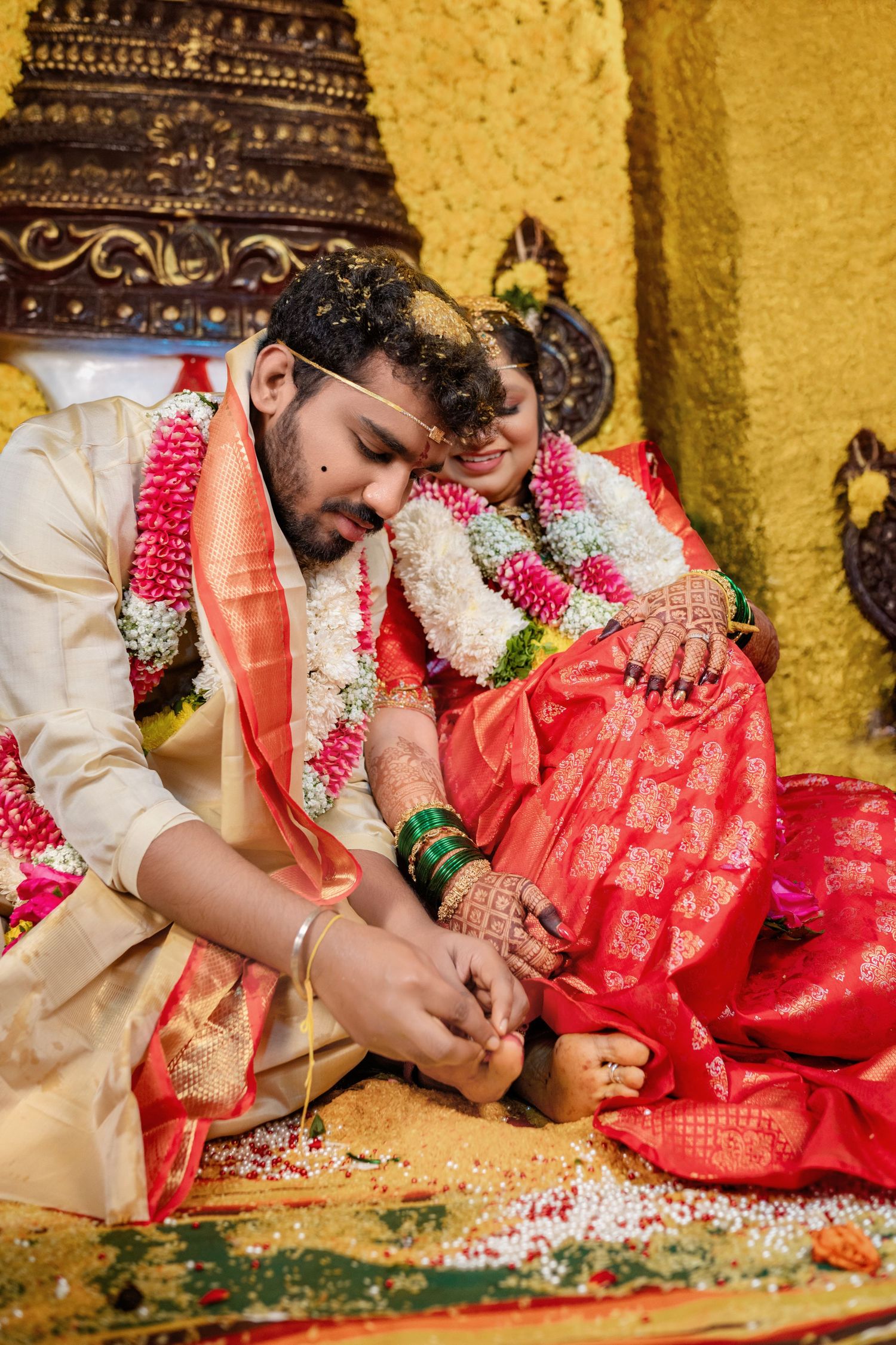 Groom placing toe ring on bride during traditional South Indian Hindu wedding ceremony with floral garlands.