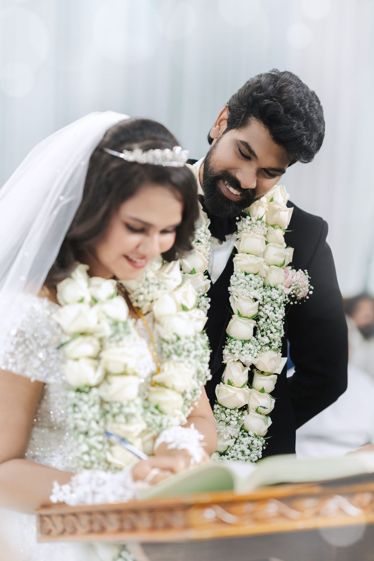 Bride and groom exchanging vows, wearing white floral garlands, signing wedding register in elegant ceremony attire.