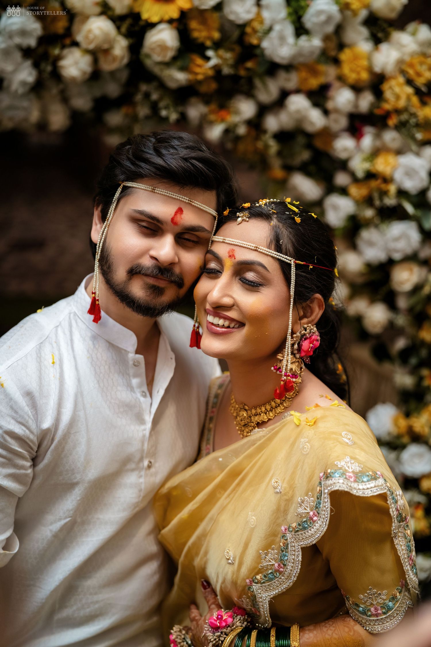 Indian couple in traditional wedding attire posing before floral backdrop, bride in yellow saree with gold jewelry.