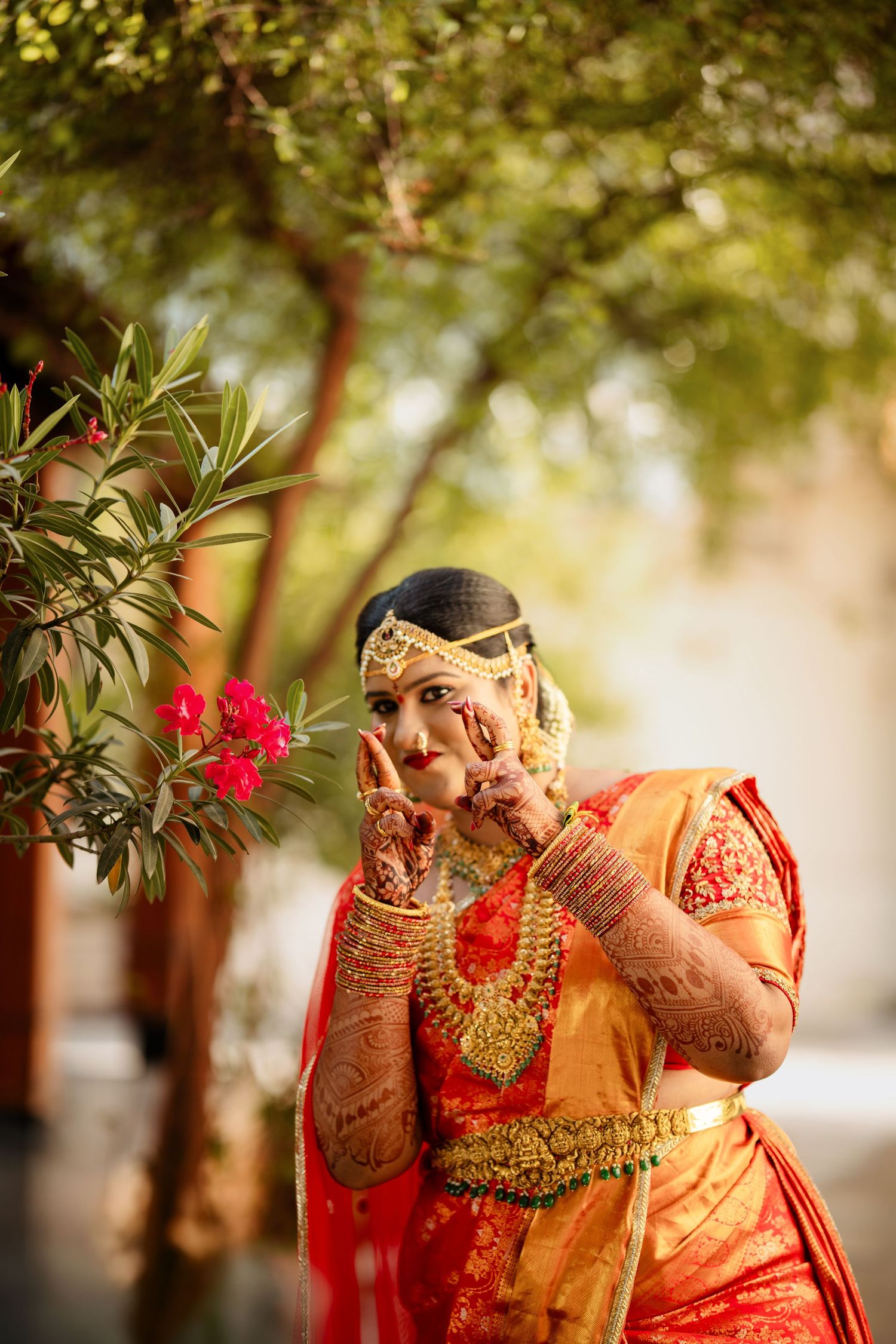 Indian bride in red and gold saree with mehndi hands and traditional jewelry poses near red flowers outdoors.