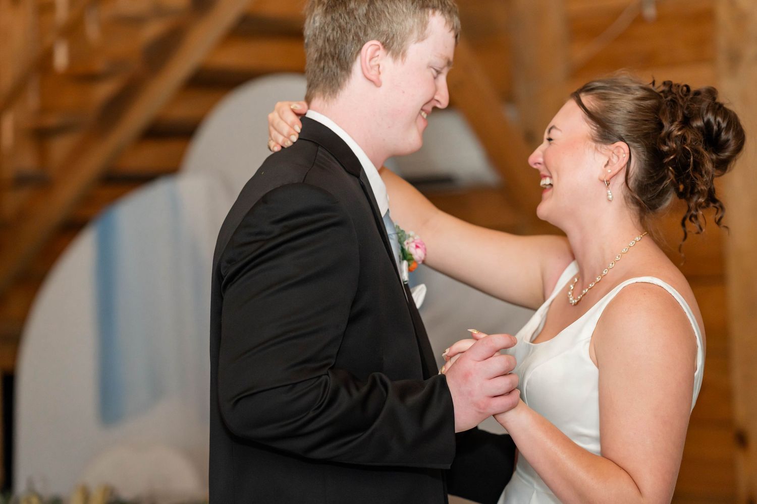 Bride and groom share their first dance, smiling joyfully, in a warmly lit wooden barn wedding venue.