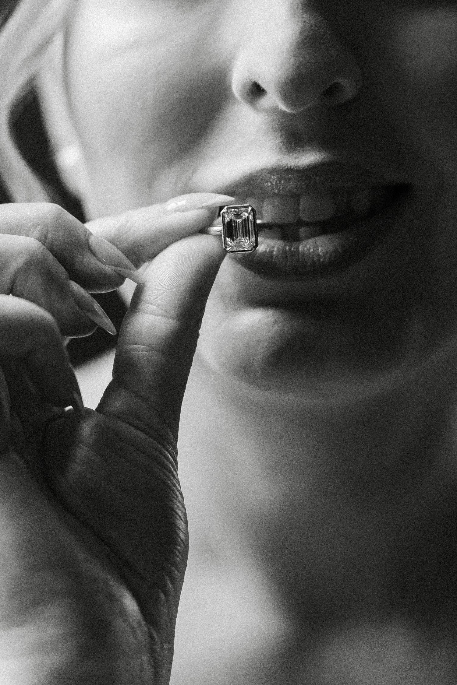Black and white close-up of a bride holding a bezel diamond engagement ring near her lips.