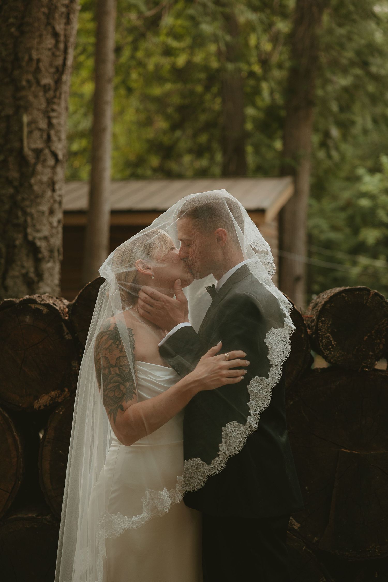 A bride and groom share a romantic kiss under a lace-trimmed veil in a forest wedding setting.