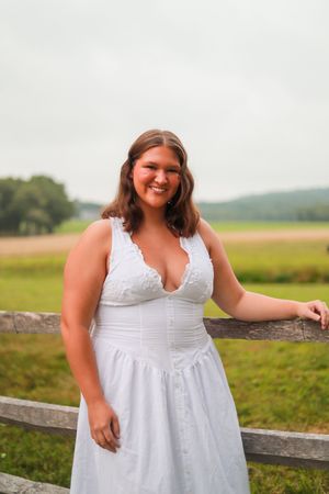 Person in white dress stands by wooden fence in rural field setting.