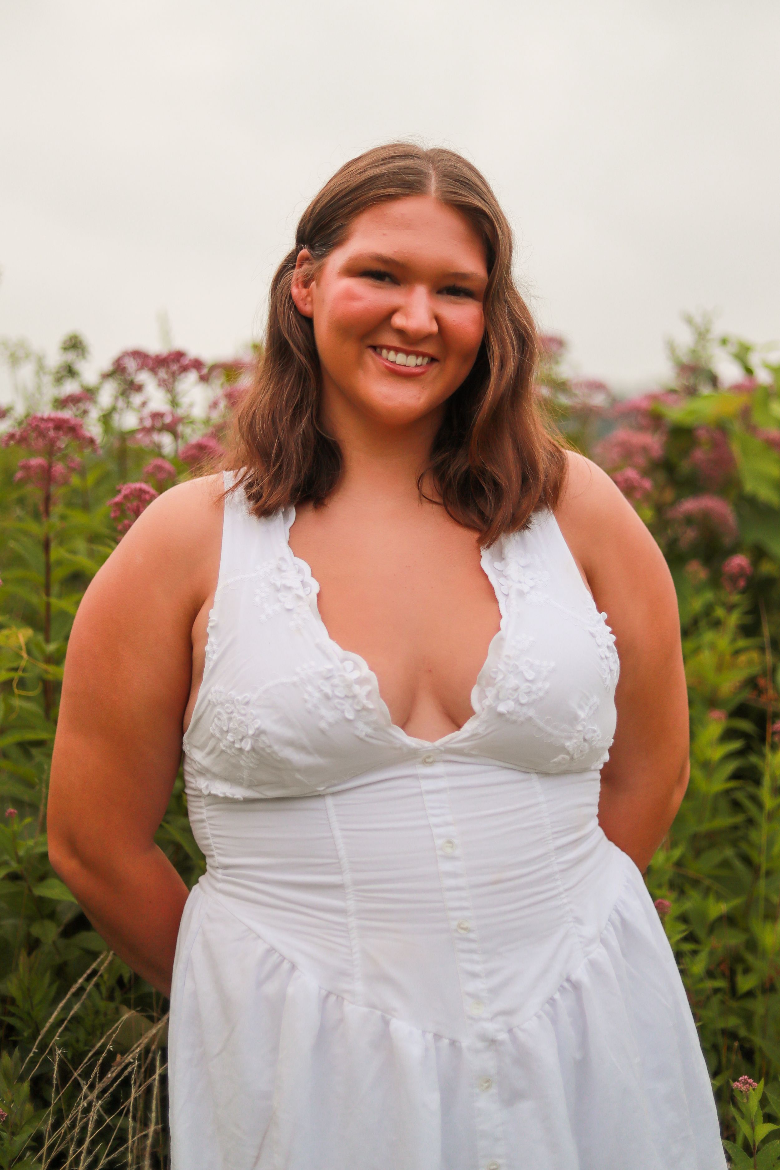 Person wearing white lace-trimmed dress poses outdoors near flowering plants.