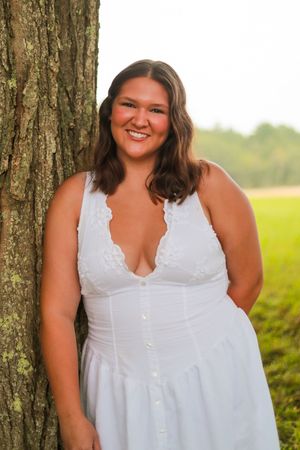 Person in white button-front dress poses near tree trunk in natural setting.