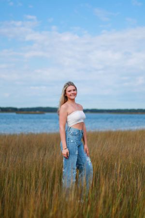 Woman wearing denim overalls stands in tall grass with ocean view behind.