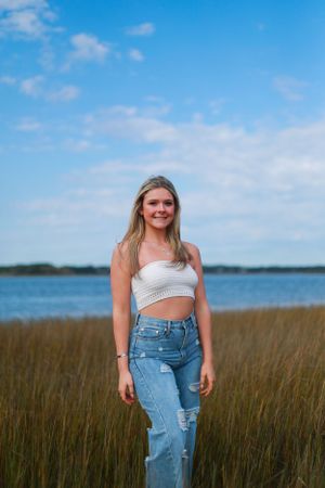 A summer portrait in tall grass with someone wearing ripped jeans and a crop top near the water.