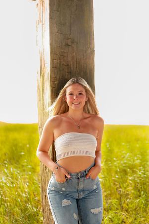A summer portrait with someone in distressed jeans and crop top standing by a wooden post in tall grass.