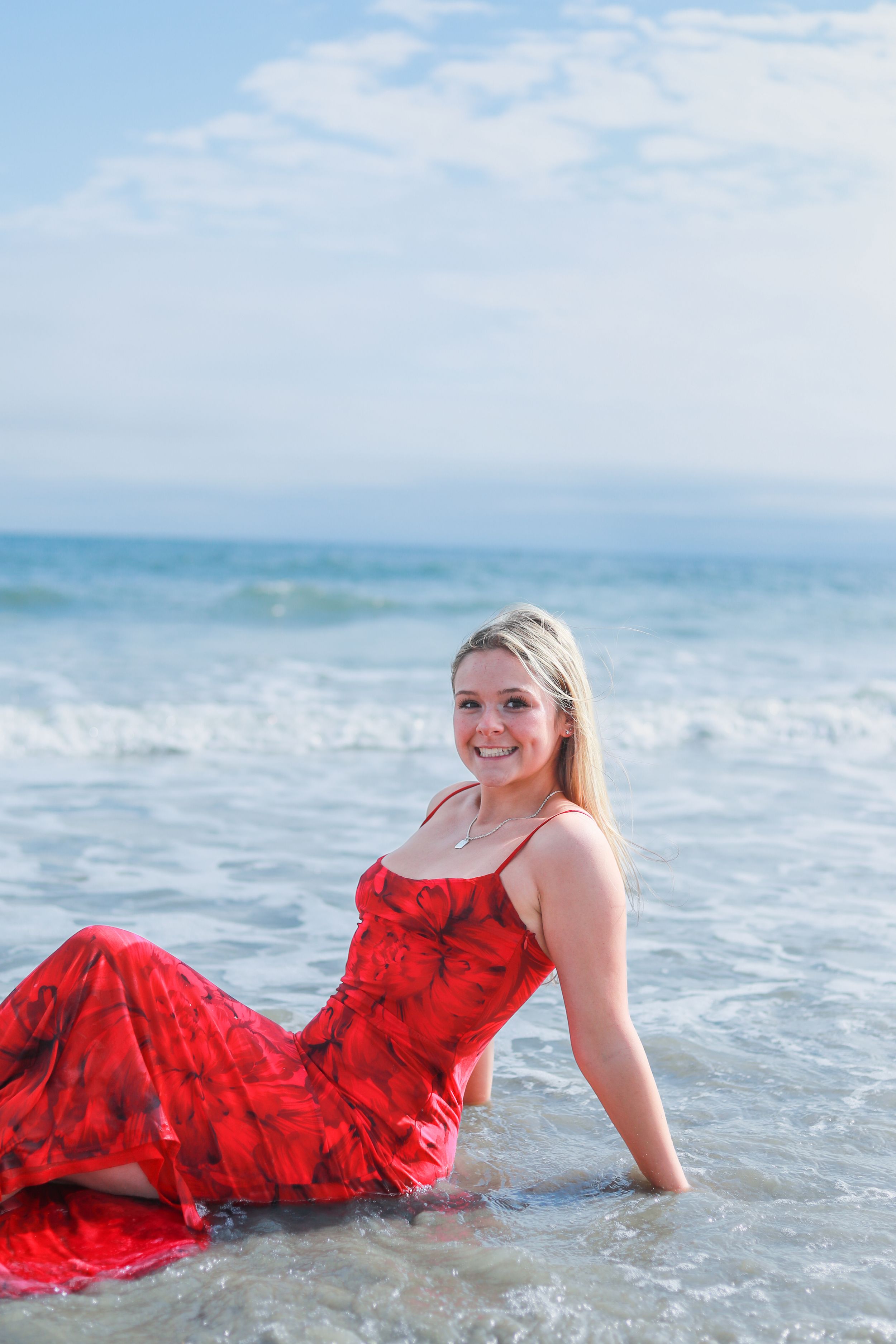 Woman in flowing red dress poses gracefully in ocean waves at beach during sunset photoshoot.