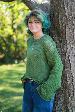 Person wearing green knit sweater and jeans poses against tree trunk in sunlit forest.