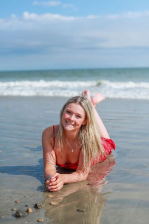 Woman in red dress lying on wet sand as ocean waves roll in during beach photoshoot.