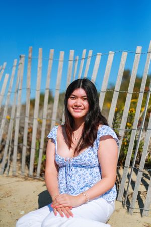 Person in blue floral dress sits by wooden beach fence on sunny day.