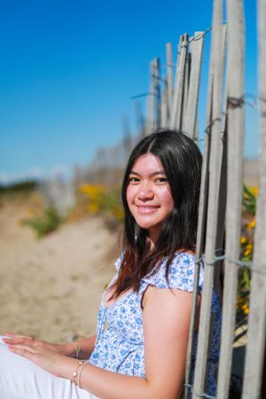 Person in blue floral dress smiles while posing near wooden beach fence on sunny day.