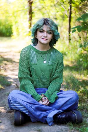 Person in green sweater and jeans sits cross-legged on forest path.