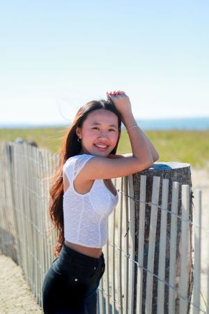 A person in a white crop top poses casually against a weathered beach fence on a sunny day.