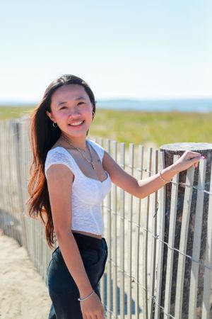 Person in white top poses playfully by beach fence on sunny day.