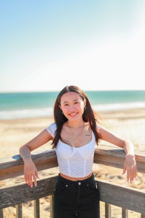 A beachside portrait showing someone in a white top leaning on a wooden railing with ocean waves behind.