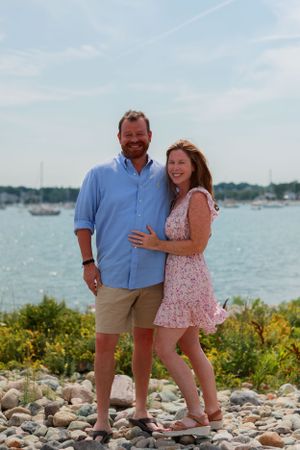 Couple enjoying time together on rocky beach with boats visible in harbor.