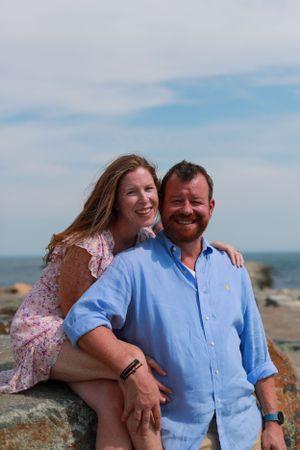 Couple poses together on beach, wearing blue shirt and floral dress.