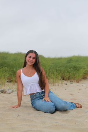Person in white top and jeans poses casually on sandy beach with dune grass.