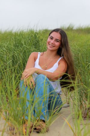 Person in white tank top and jeans relaxes among beach grass during outdoor photoshoot.