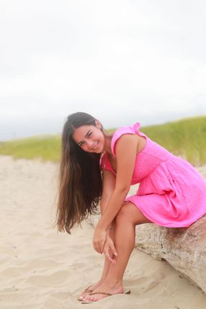 Person in pink sundress poses playfully on sandy beach during summer photoshoot.