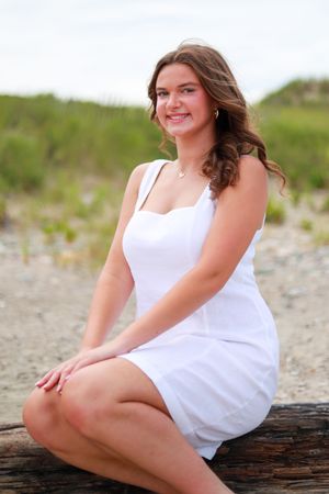 Portrait of person in white dress seated on driftwood at beach.