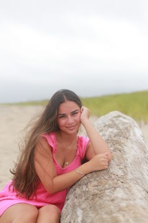 A person in a pink dress poses casually against a large rock at the beach on an overcast day.