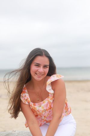A person in a floral top and white pants smiles while posing on a sandy beach.