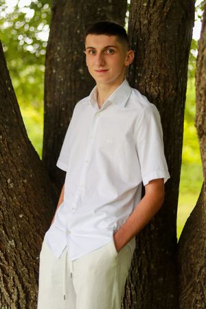 A person in white shirt and light pants poses between tree trunks in a park setting.