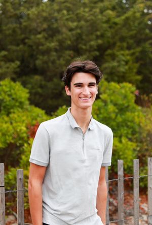 Person in white polo shirt poses near wooden fence with greenery in background.