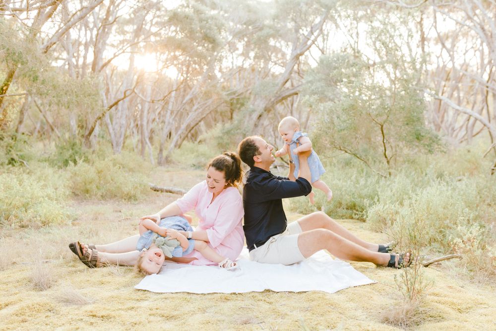 Family sitting on white blanket outdoors at golden hour, parents with two young children in soft natural light.