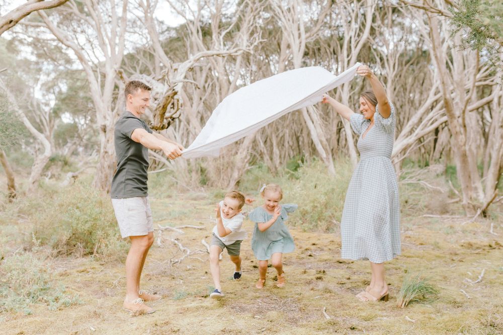 Family of four playing with a large white umbrella outdoors among gum trees on a sunny day.