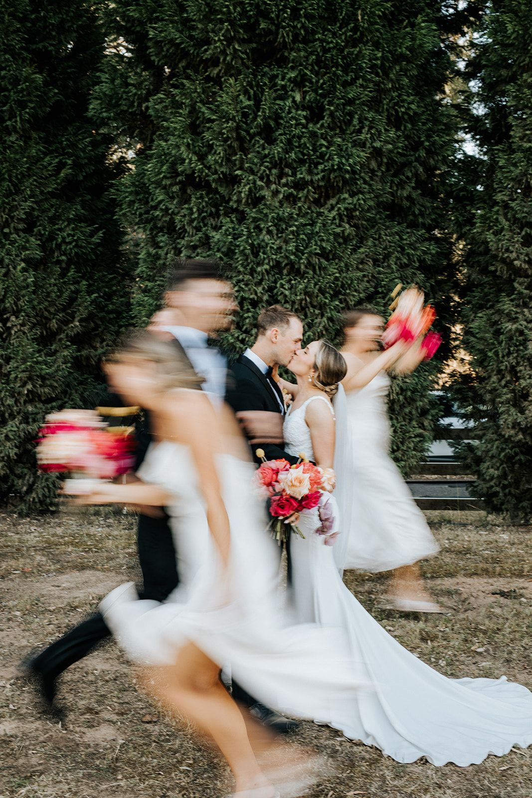Bride and groom kissing at wedding while bridal party runs past with pink bouquets, motion blur effect, green trees background.