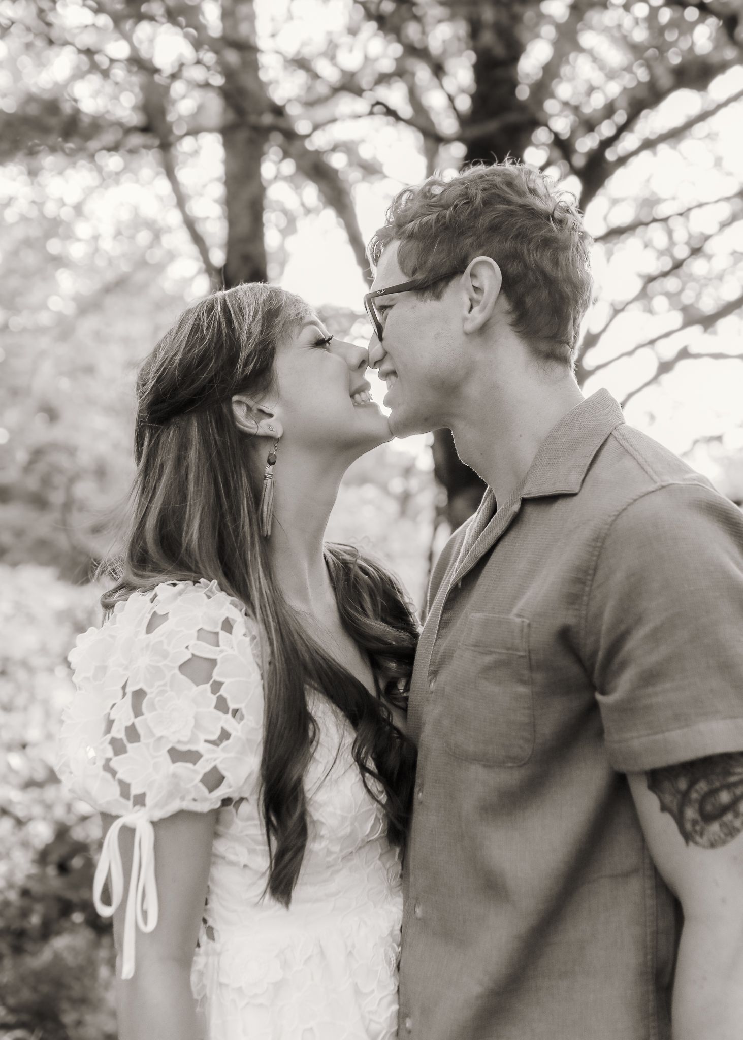 Black and white photo of a couple sharing a romantic moment, nearly kissing outdoors under trees.