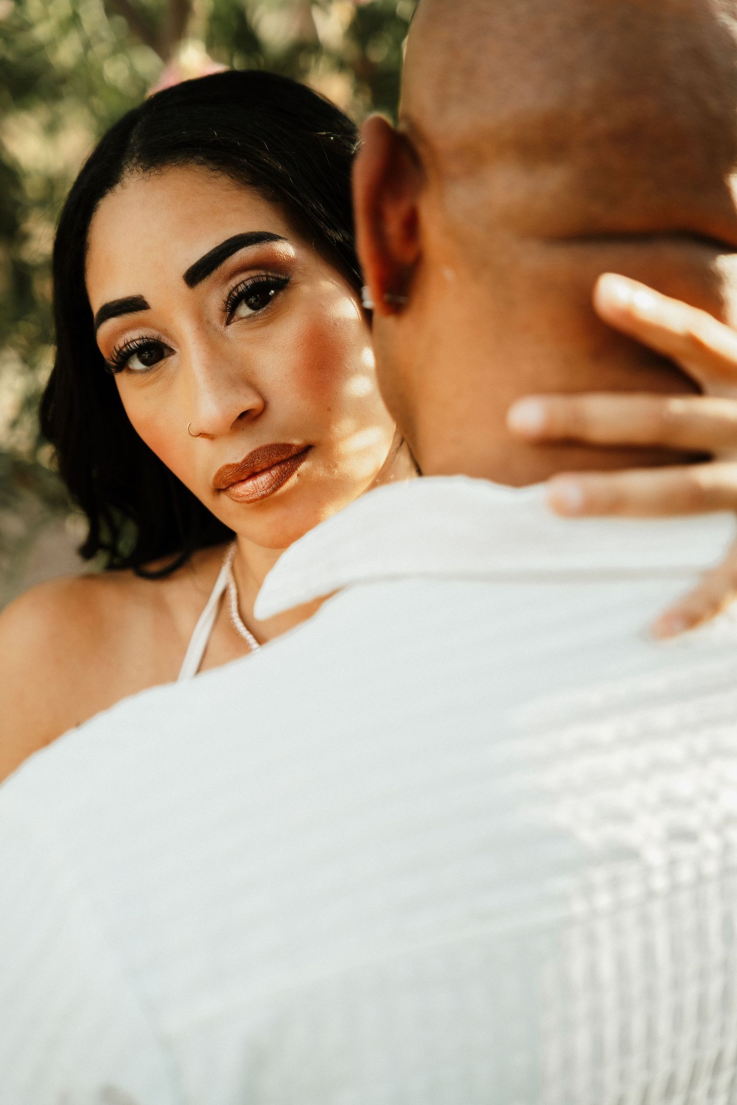 A woman with dark hair and bold makeup gazes over a man's shoulder during a romantic outdoor couples portrait session.