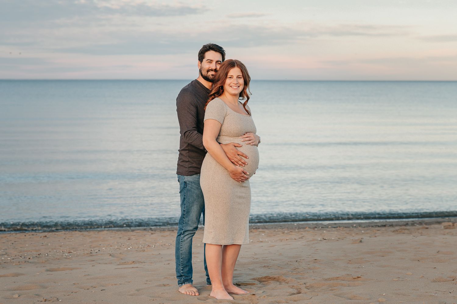Pregnant couple posing on sandy beach at sunset, man embracing woman in beige maternity dress near calm ocean waters.