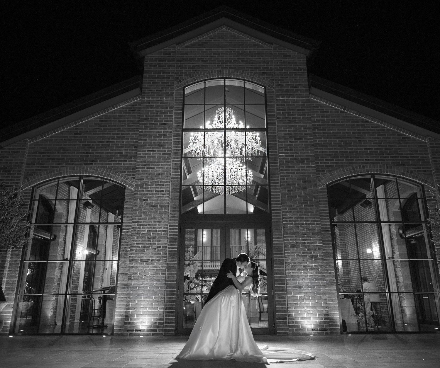Bride and groom share romantic kiss outside illuminated brick wedding venue at night in black and white photo.