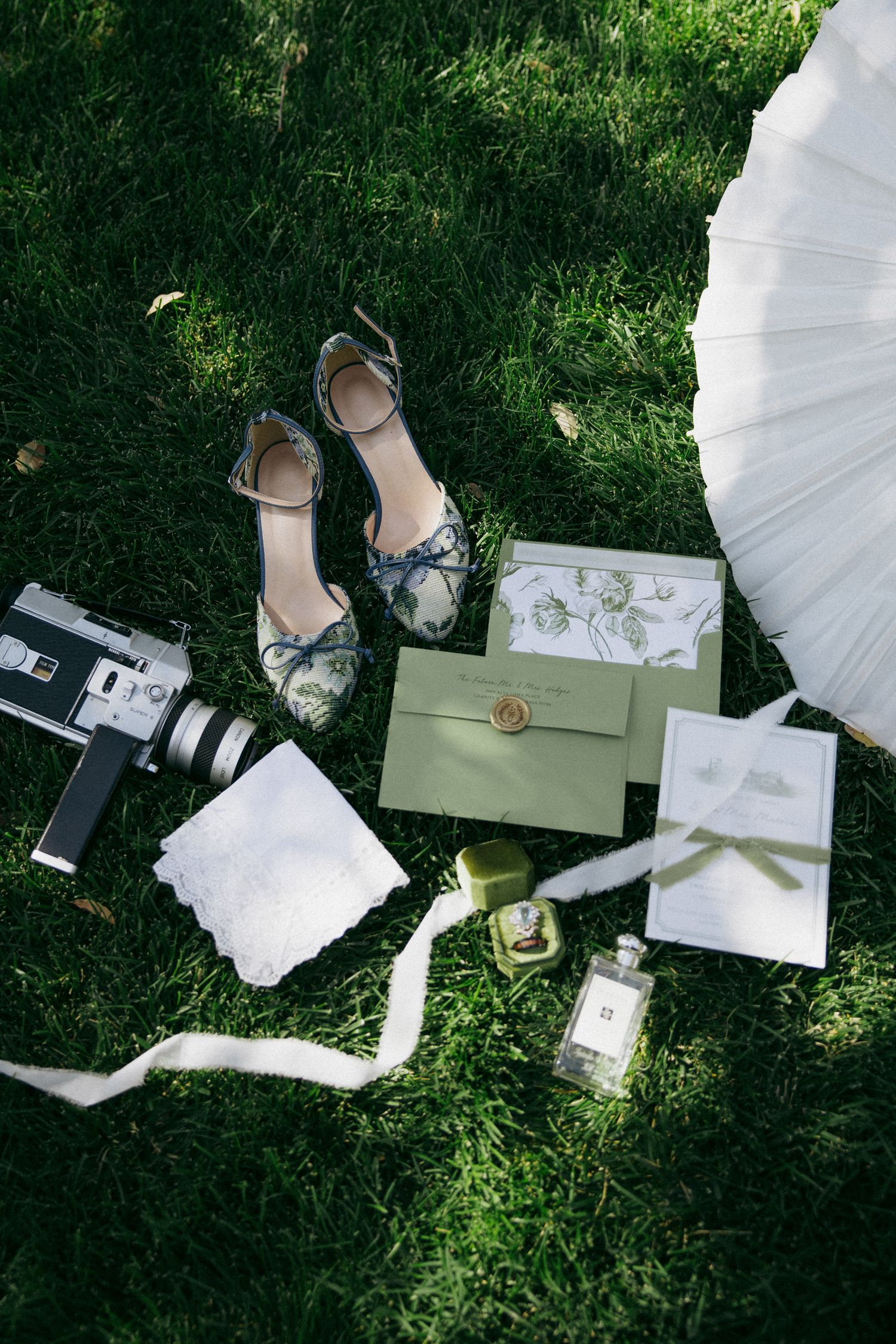 Elegant wedding flat lay on grass featuring heels, camera, invitations, perfume, and parasol.