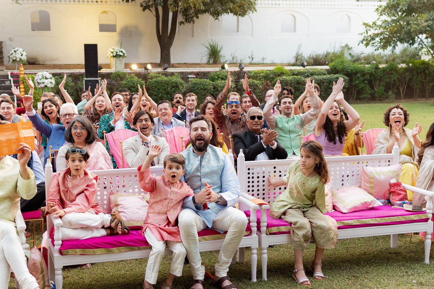 Joyful wedding guests in colorful traditional attire cheering with raised hands at an outdoor Indian celebration.
